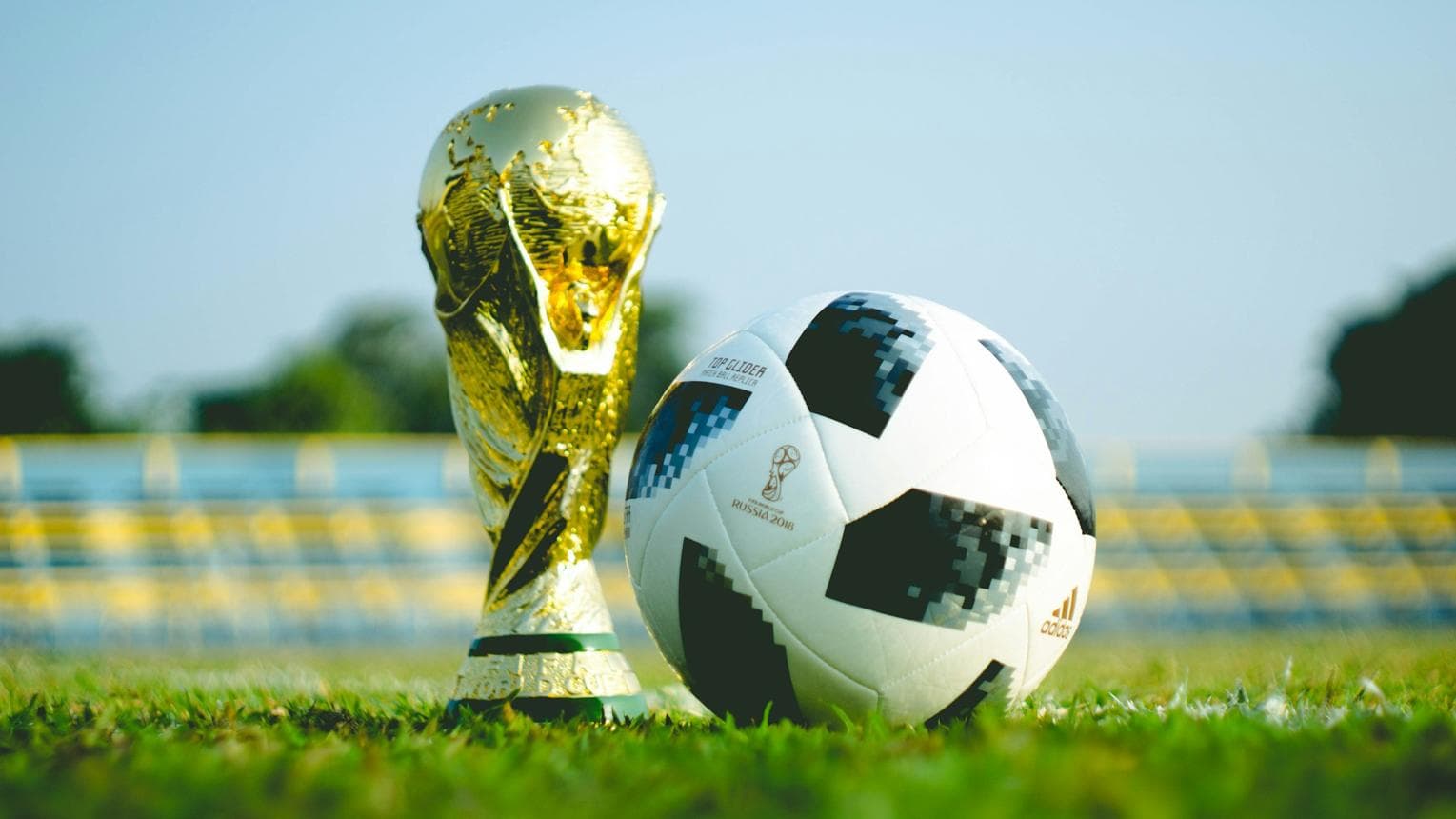 FIFA World Cup gold trophy beside a football on a grass pitch at sunset.